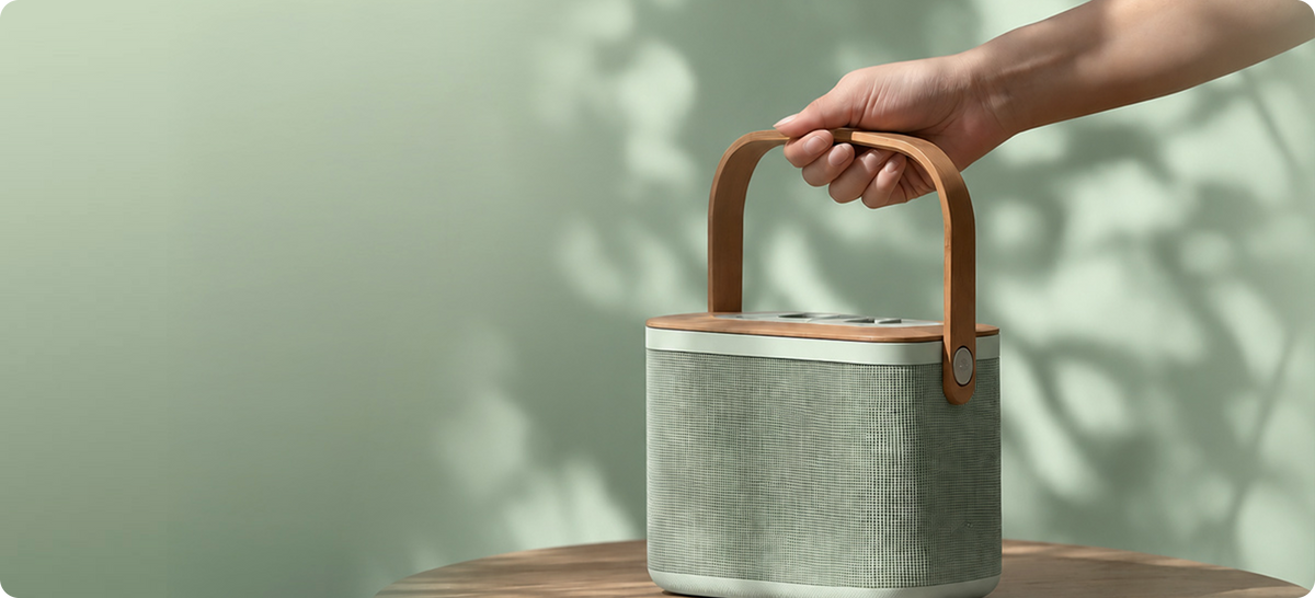 A light sage green fabric speaker with a brown leather handle, held by a person's hand on a wooden table against a soft green background with leaf shadows.