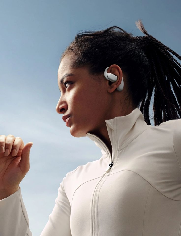 A woman in a white athletic zip-up jacket wearing white wireless earbuds, shown in profile against a clear blue sky.1