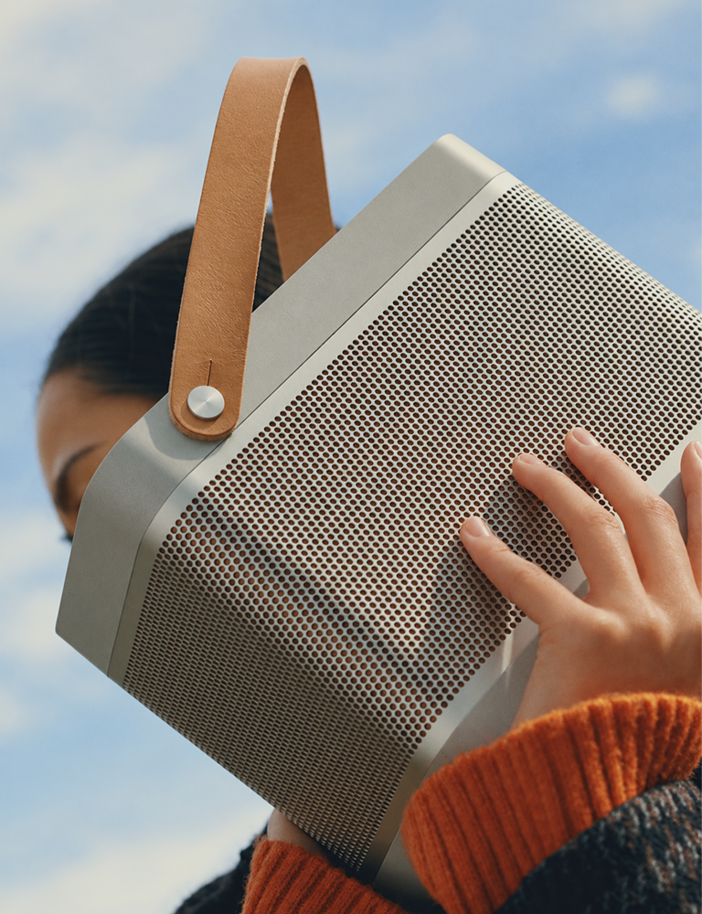 A close-up lifestyle shot of a portable Bluetooth speaker being held by a person outdoors against a blue sky. The speaker has a gray perforated metal grille, a light gray frame, a tan leather handle, and is worn over an orange knit sweater.4